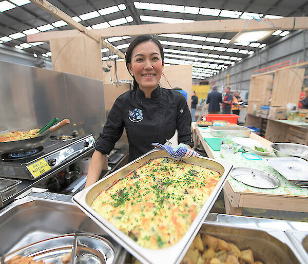 A woman in black catering costume holds up a tray of potato bake in kitchen surroundings.