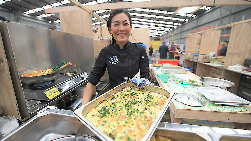 A woman in black catering costume holds up a tray of potato bake in kitchen surroundings.