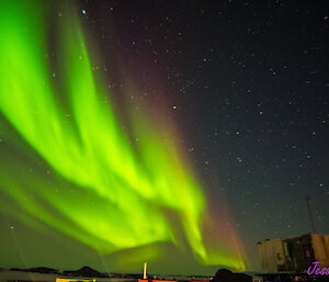 A bright green, yellow, and purple aurora is visible in the night sky above station buildings