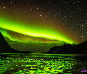 A bright green aurora is visible against the clouds in the night sky. In the foreground is a frozen lake reflecting the colours of the aurora and there are dark, rocky mountains to the left and right of frame.