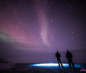 Two men aree standing on a frozen lake that is lit by light from within. A pale aurora is visible in the night sky above them.