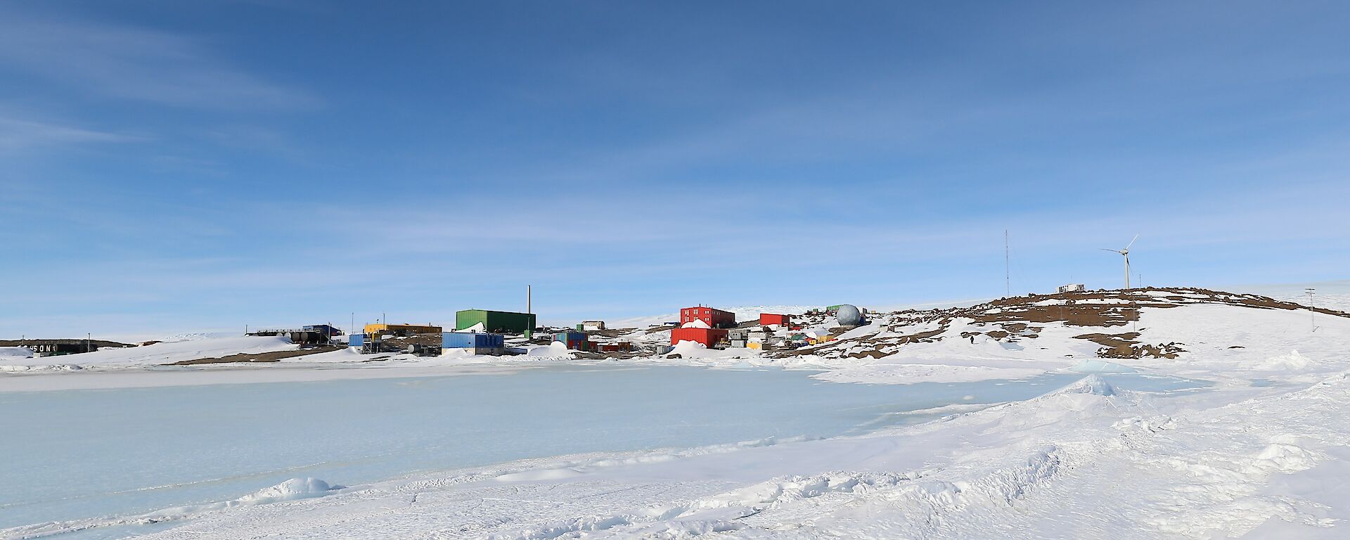Brightly coloured buildings of a station are on a rocky, ice and snow covered landscape in the distance. In the foregound is an iced over harbour