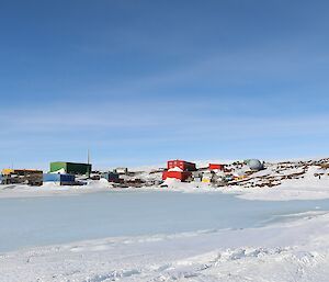 Brightly coloured buildings of a station are on a rocky, ice and snow covered landscape in the distance. In the foregound is an iced over harbour