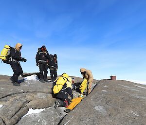Six men are gathered around a mannequin that is laying in a crack on a rocky hillside.
