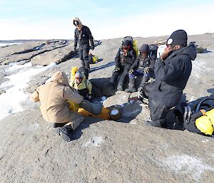Six men are providing simulated first aid to a mannequin that is laying on a rocky hillside.