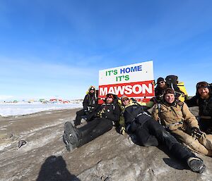 Six men are sitting next to a sign that says "It's Home It's Mawson" . IIn the distance is a group of brightly coloured buildings on an ice covered rocky landscape.