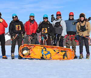 Seven men are posing in line for a photograph on an ice plateau. At their feet is a mannequin strapped securely to a lifting stretcher.