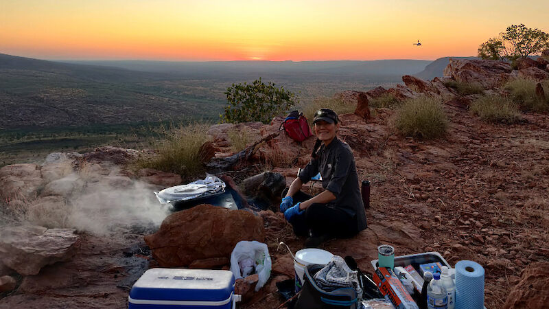 A woman smiling, sitting on a cliff at dawn, surrounded by eskies and supplies