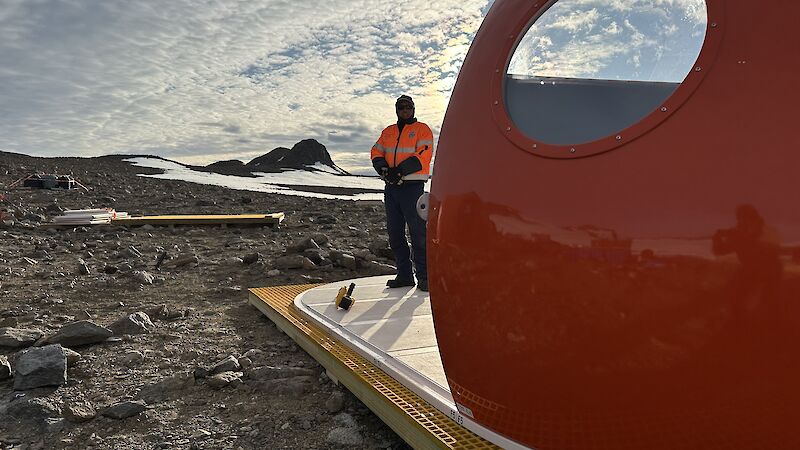 A man in orange high vis stands near an orange dome tent with rocks and ice behind him