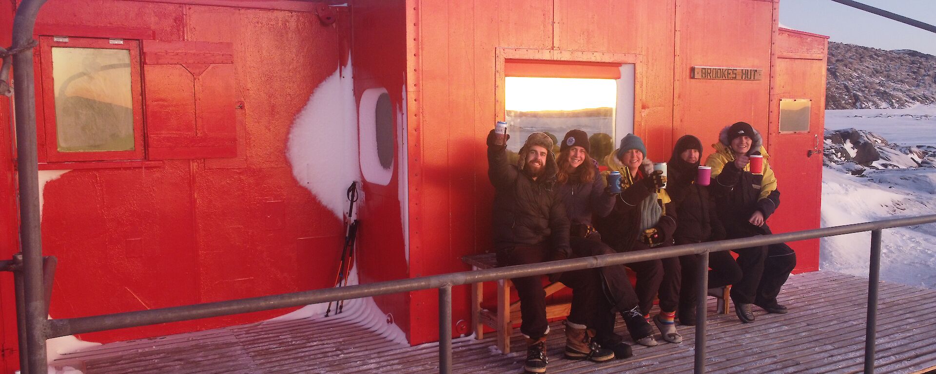 Five smiling people sitting on a bench out the front of a red hut.