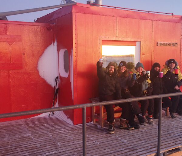 Five smiling people sitting on a bench out the front of a red hut.