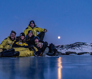 Six smiling people posing for a photo on the sea ice with the rising moon behind them over snow covered hills.