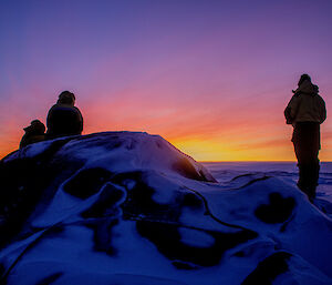 Three people standing atop a snow-covered hill look into the distance to a rising sun that is streaking the sky yellow and orange.