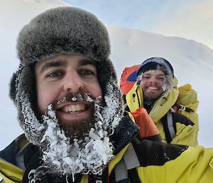 Two smiling men wearing snow gear, one whose beard is covered in icicles and frost from the snow.