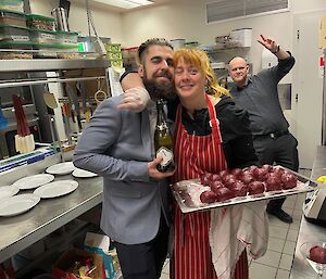 A kitchen scene: a man in a suit hugs a lady wearing a striped apron and holding a tray of food while another man waves in the background.