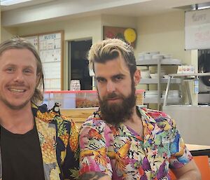 Two smiling men wearing bright colourful shirts with a kitchen visible behind them.