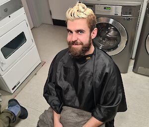 A smiling man sitting on a chair wearing a protective shawl has just had the top of his hair dyed platinum blonde.