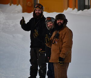 Three men in warm winter clothing standing in a line and smiling at the camera. The background is snow covered ground.