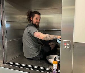 A smiling man sits inside a stainless-steel dumb waiter and cleans the surfaces.
