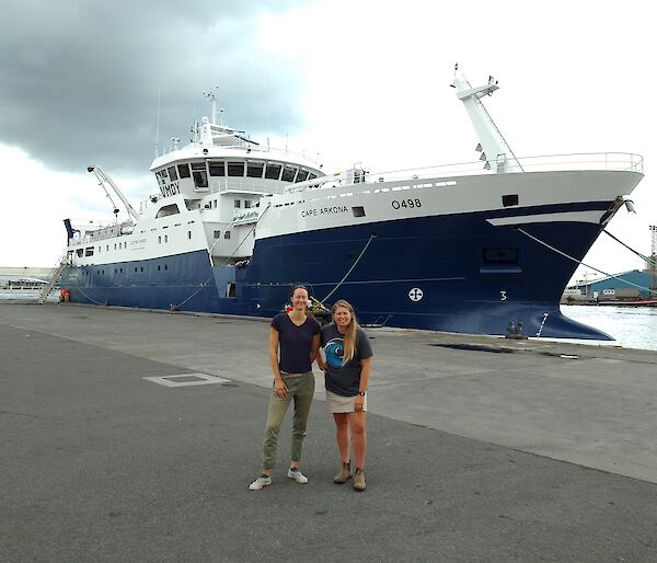 Two women standing in front of a large blue and white fishing vessel, tied up on a cement wharf