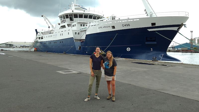 Two women standing in front of a large blue and white fishing vessel, tied up on a cement wharf