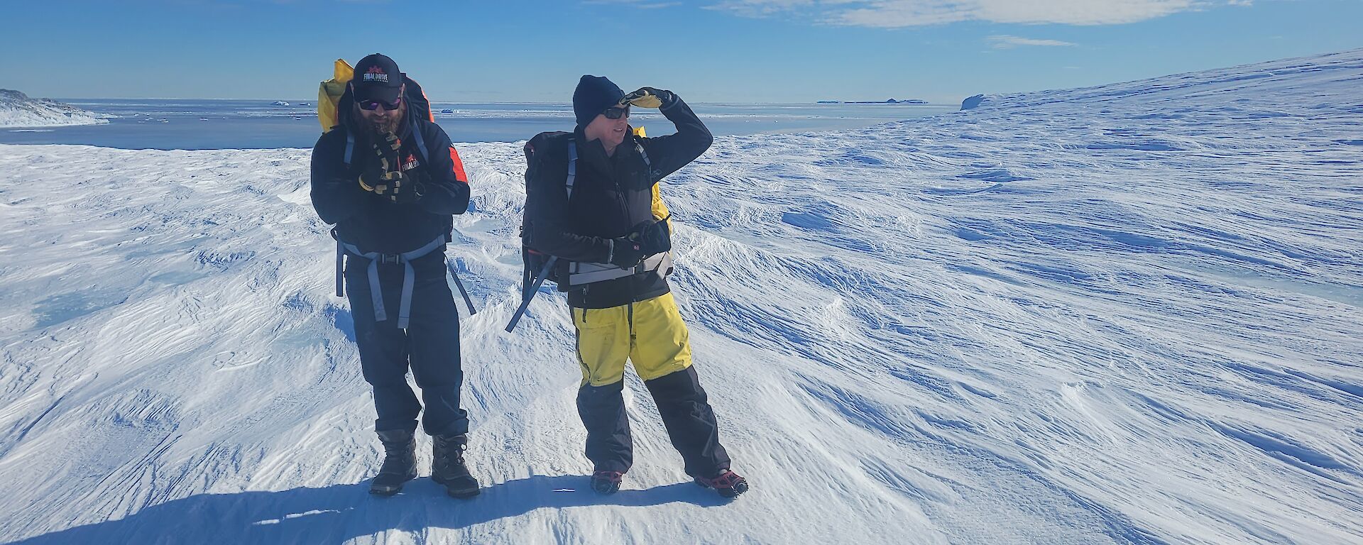 Two Antarctic expeditioners stand on sastrugi on a sunny day.
