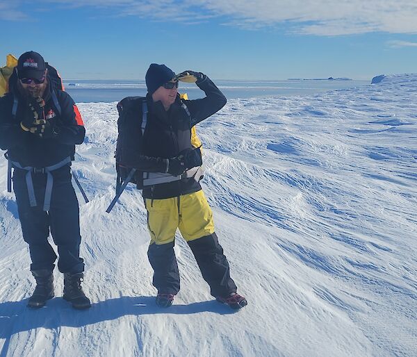 Two Antarctic expeditioners stand on sastrugi on a sunny day.