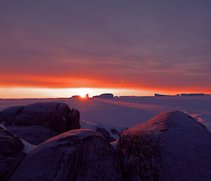 A radiant, orange, sunset is visible on the horizon highlighting icebergs in the distance on the sea ice. In the foreground is a rocky island landscape.