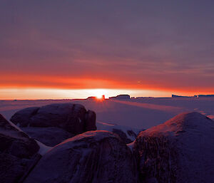 A radiant, orange, sunset is visible on the horizon highlighting icebergs in the distance on the sea ice. In the foreground is a rocky island landscape.