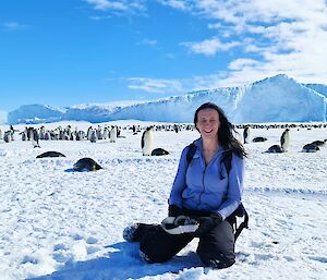 A woman is smiling and looking at the camera as she kneels on the sea ice. Behind her is a large number of emperor penguins and on the horizon are large icebergs rising from the sea ice.