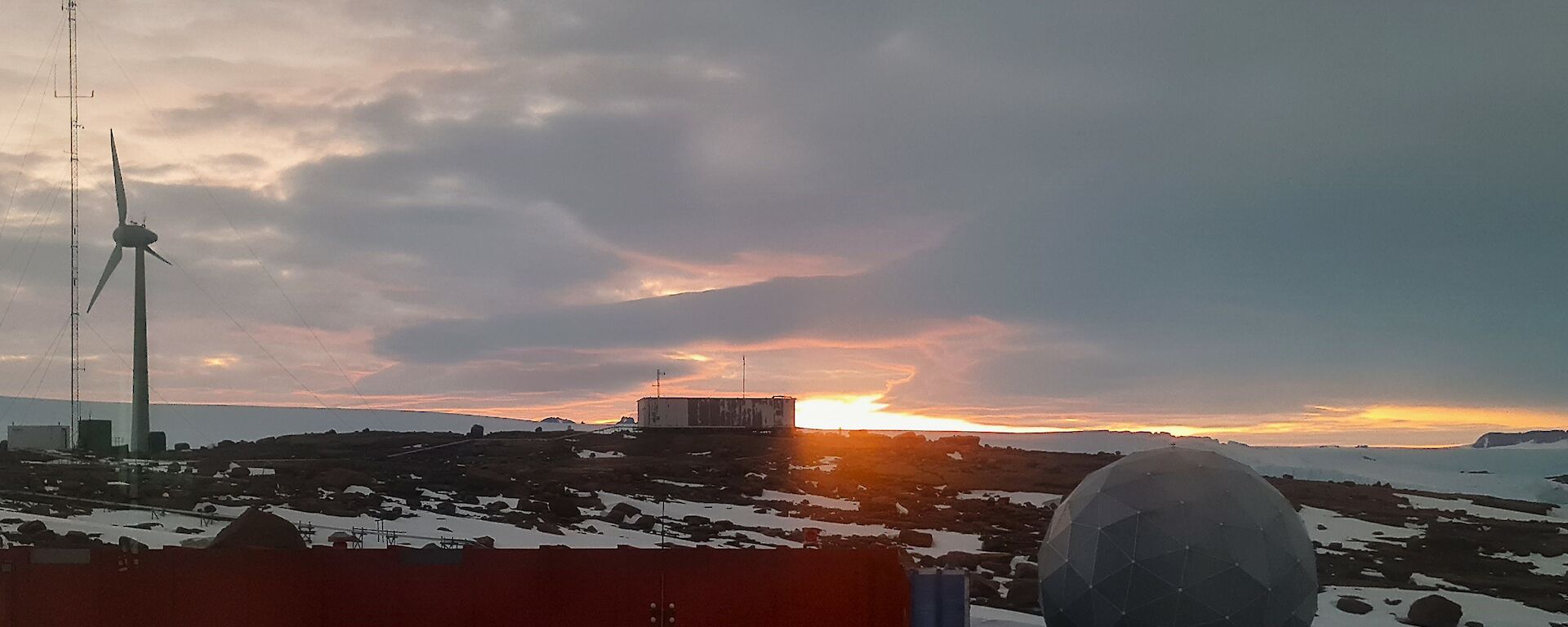 The orange glow of the sun is visible on the horizon beyond an old building that sits atop a rocky, snow and ice covered hill. There is a large wind turbine to the left of frame and a satellite dome to the bottom right of frame.