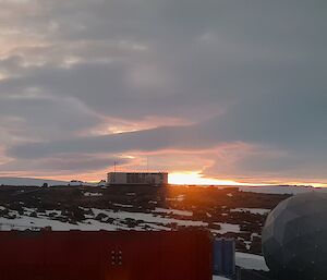 The orange glow of the sun is visible on the horizon beyond an old building that sits atop a rocky, snow and ice covered hill. There is a large wind turbine to the left of frame and a satellite dome to the bottom right of frame.