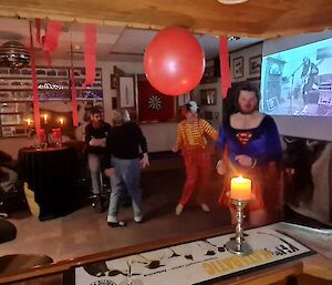 A number of people are dancing in a bar area. Some are dressed in halloween costumes and there are red streamers and a balloon hanging from the ceiling. A video screen is on in the background.