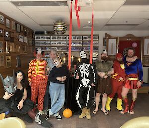 A group of people aree gathered for a photograph. They are dressed in a variety of halloween costumes and there are red streamers hanging from the ceiling.