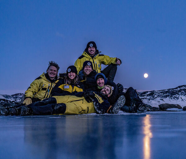 A group of expeditioners in yellow and black sit by a frozen lake with a full moon behind.