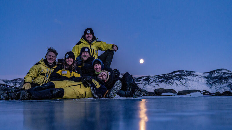 A group of expeditioners in yellow and black sit by a frozen lake with a full moon behind.