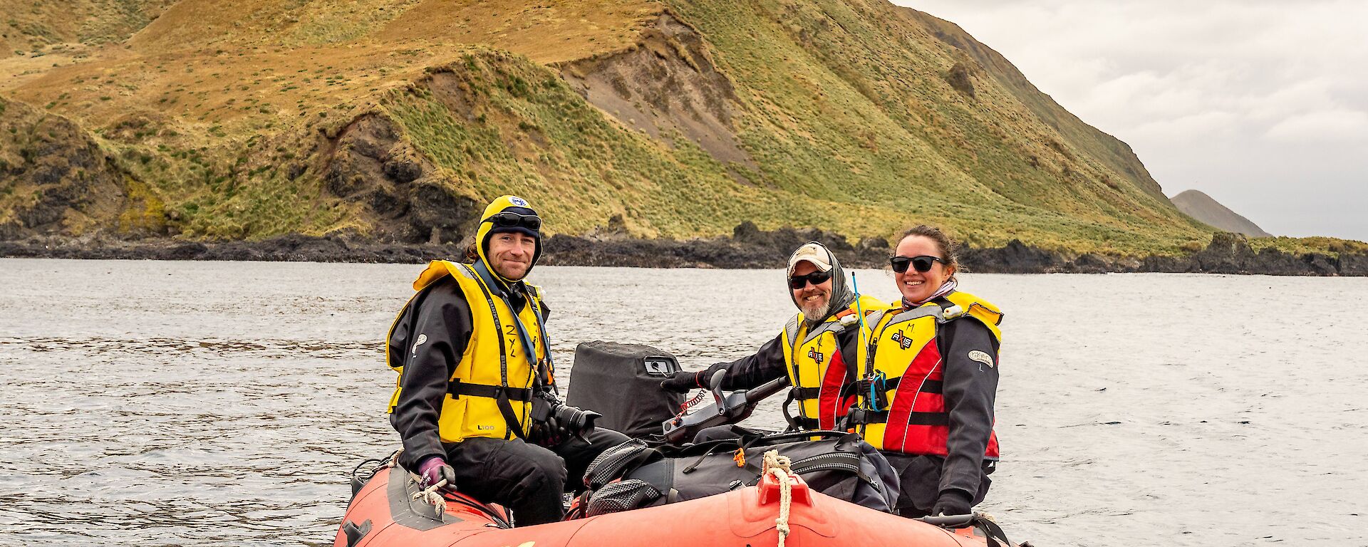 Three people in life jackets sit in a red dinghy with a barren hill behind them