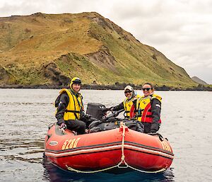 Three people in life jackets sit in a red dinghy with a barren hill behind them