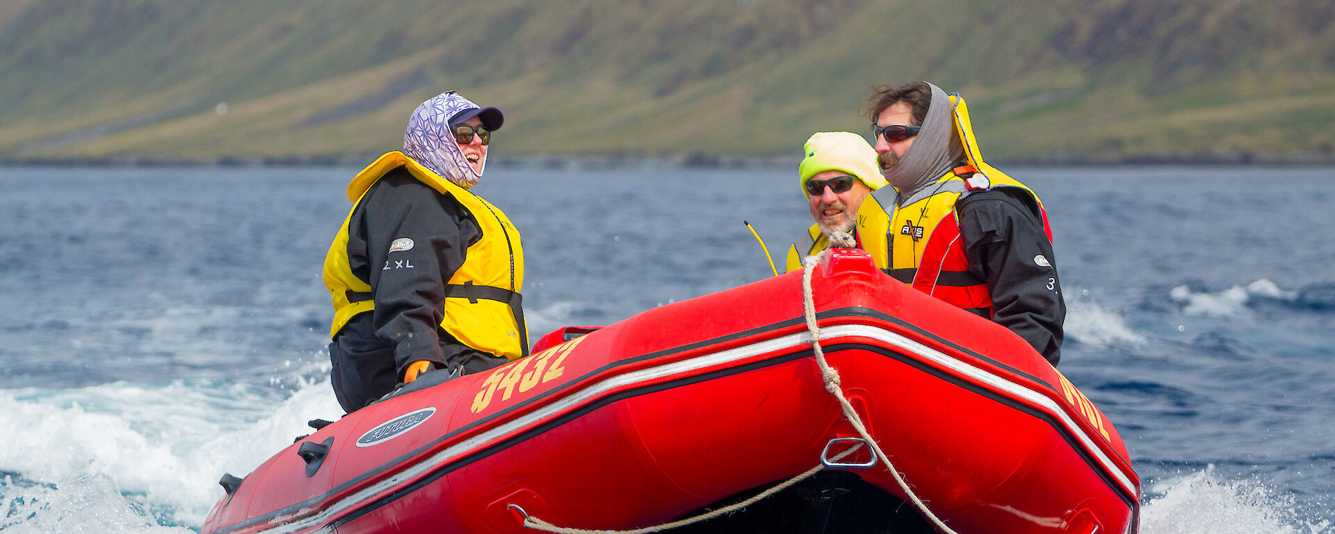 Red dinghy speeding along the water with three people in the boat and steep unwooded hills behind them