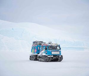 A blue Hagglunds vehicle on the sea-ice dwarfed by a large iceberg behind it.