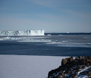 In the distance the leading edge of a glacier juts into an icy sea. In the foreground frozen sea-ice and a rocky outcrop.