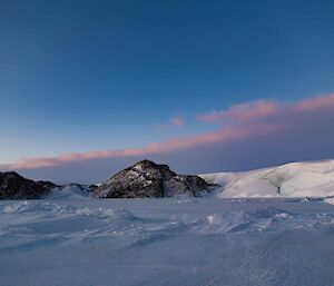 Pinkish coloured clouds above rocky snowy hills and frozen sea-ice in the foreground with lumpy areas of snow accumulation.