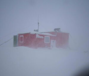 A mostly red hut with some green shrouded in misty snow during poor weather conditions.