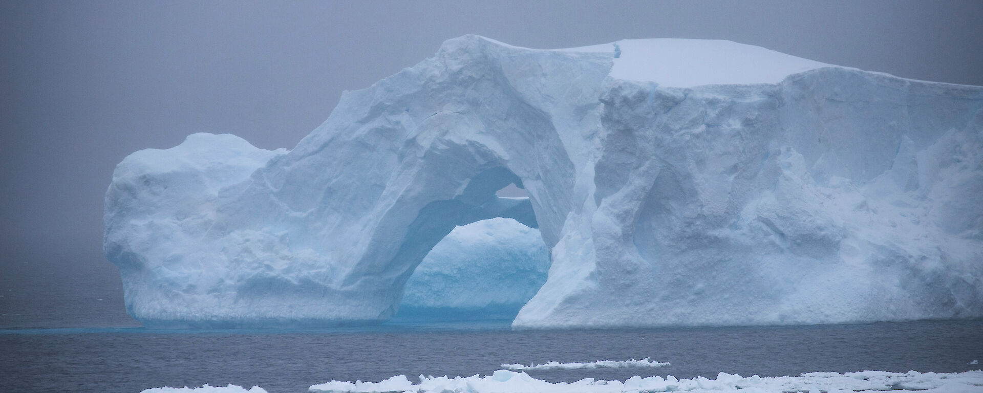 An iceberg shaped like a bridge with a hollow entrance can be seen on an overrcast day