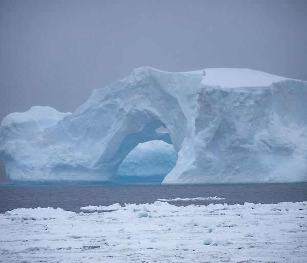 An iceberg shaped like a bridge with a hollow entrance can be seen on an overrcast day