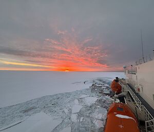The sun is setting over the sea ice. On the right of frame the side of a ship can be seen with two emergency lifeboats visible.
