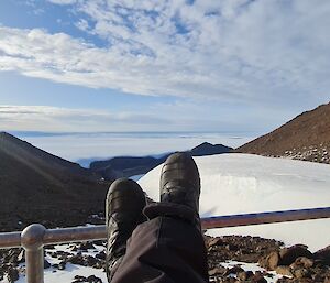 A view across to the horizon over a snow and ice covered landscape with rocky mountain ridges rising to the left and right of frame. In the foreground a woman wearing boots is resting her feet on a railing
