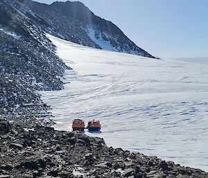 An orange Hägglunds vehicle is driving onto the ice plateau from a rocky mountainous area.