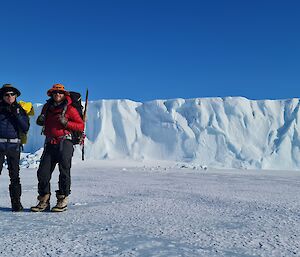 Two men in hiking equipment and carrying packs are standing on the sea ice. In the background is a large ice berg