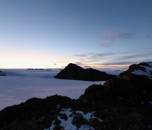 The glow of the sun below the horizon is lighting up an ice covered lanscape punctuated by rocky mountains